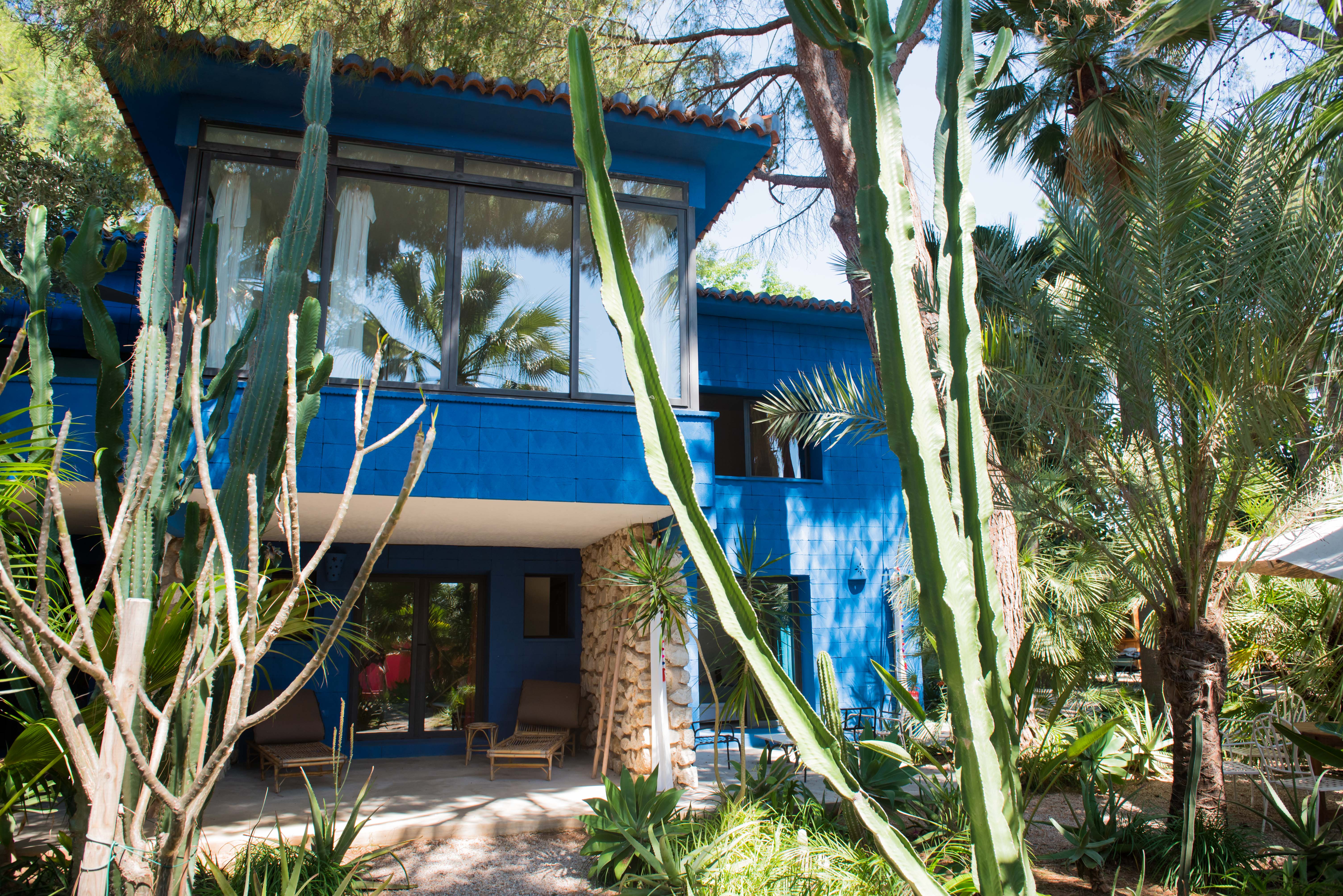 La maison bleue à deux étages offre un patio ombragé au rez-de-chaussée avec chaises longues en bambou et piliers en pierre. Le jardin tropical est encadré par des cactus et palmiers.