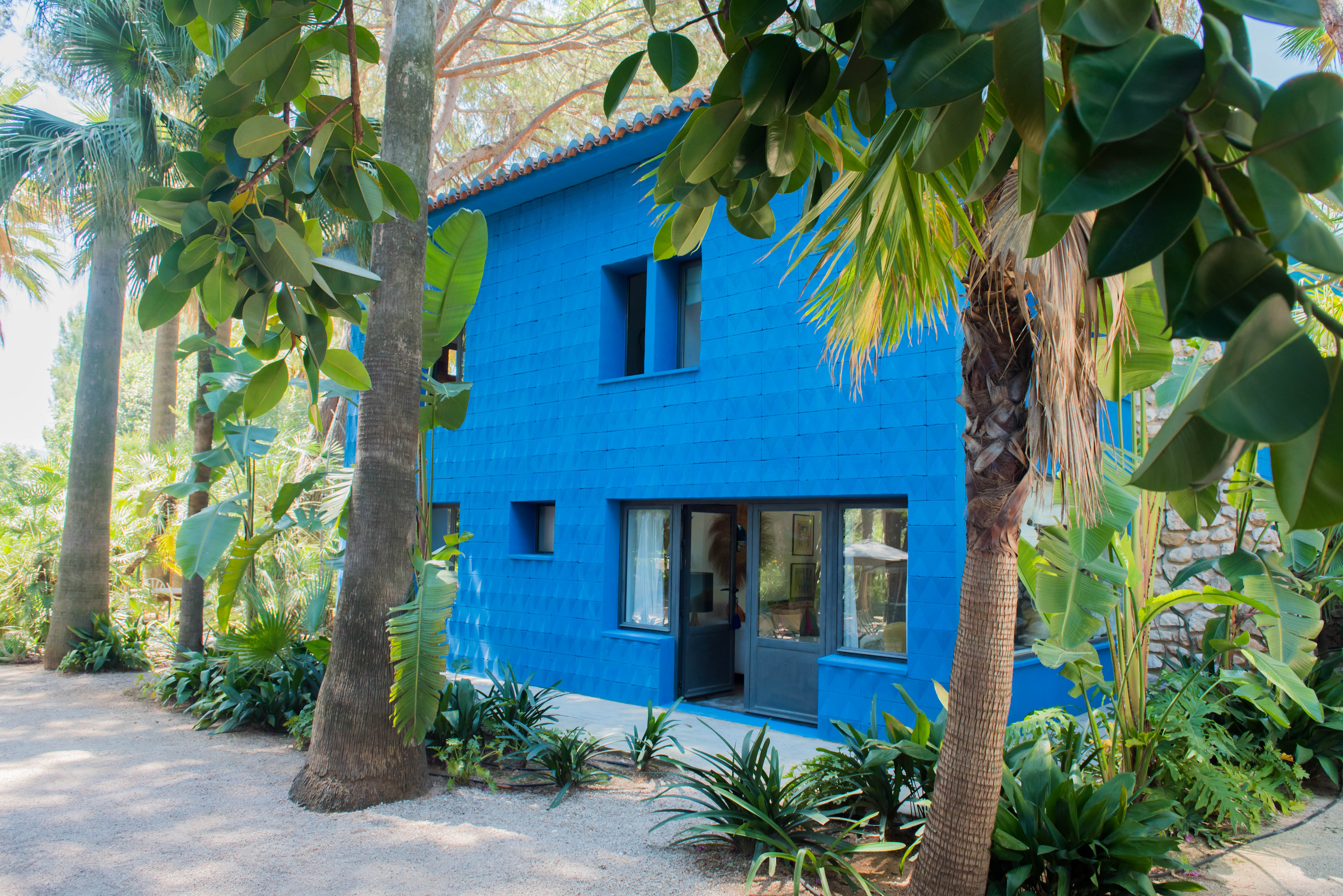 Maison à deux étages avec une façade bleue vive texturée et un toit de tuiles en terre cuite, nichée dans un jardin tropical luxuriant avec des palmiers et un chemin de gravier.
