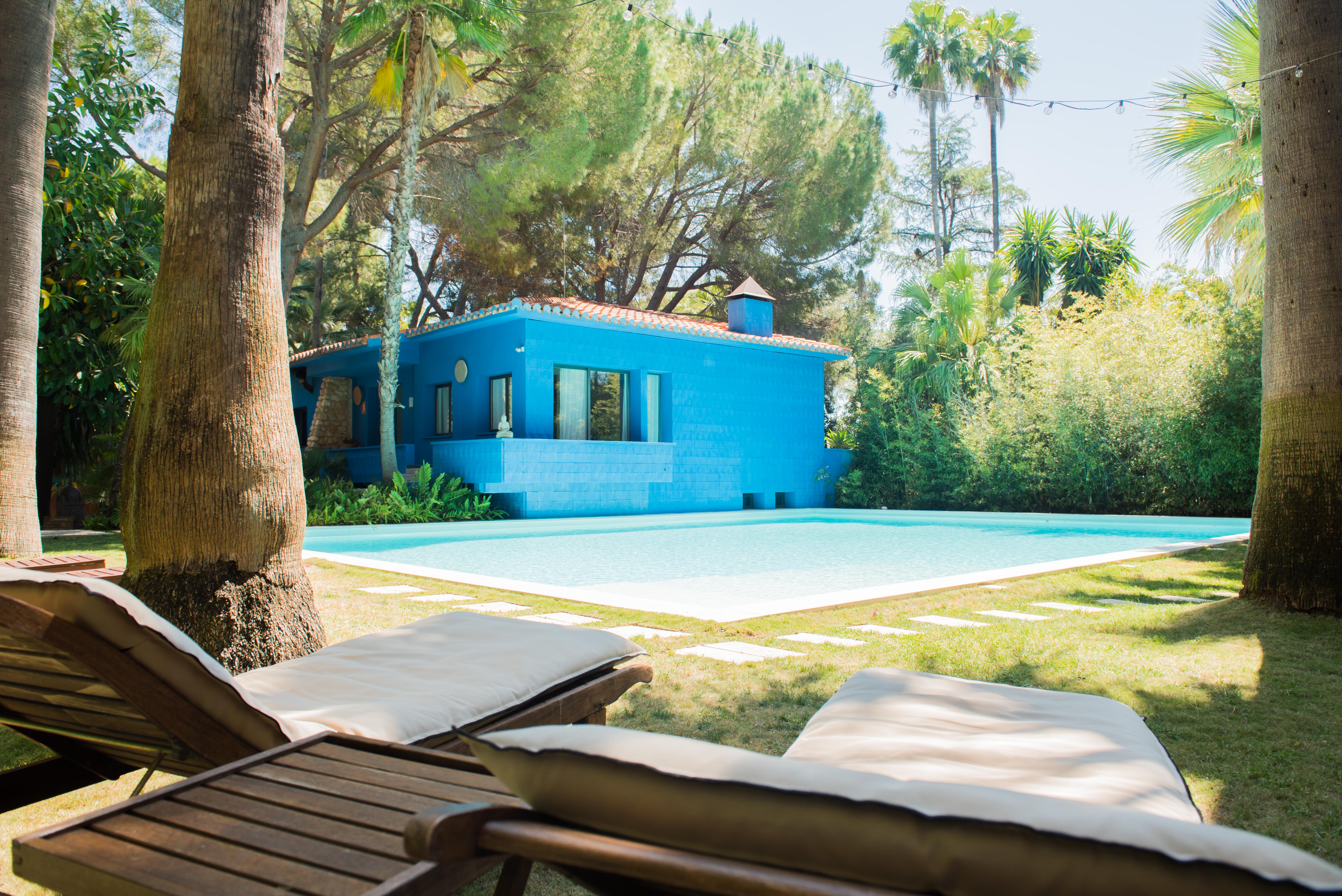 Maison bleu vif à toit de tuiles, bordant une piscine claire. Deux chaises longues en bois sur la pelouse soignée, encadrées par des palmiers et une végétation luxuriante sous des guirlandes lumineuses.