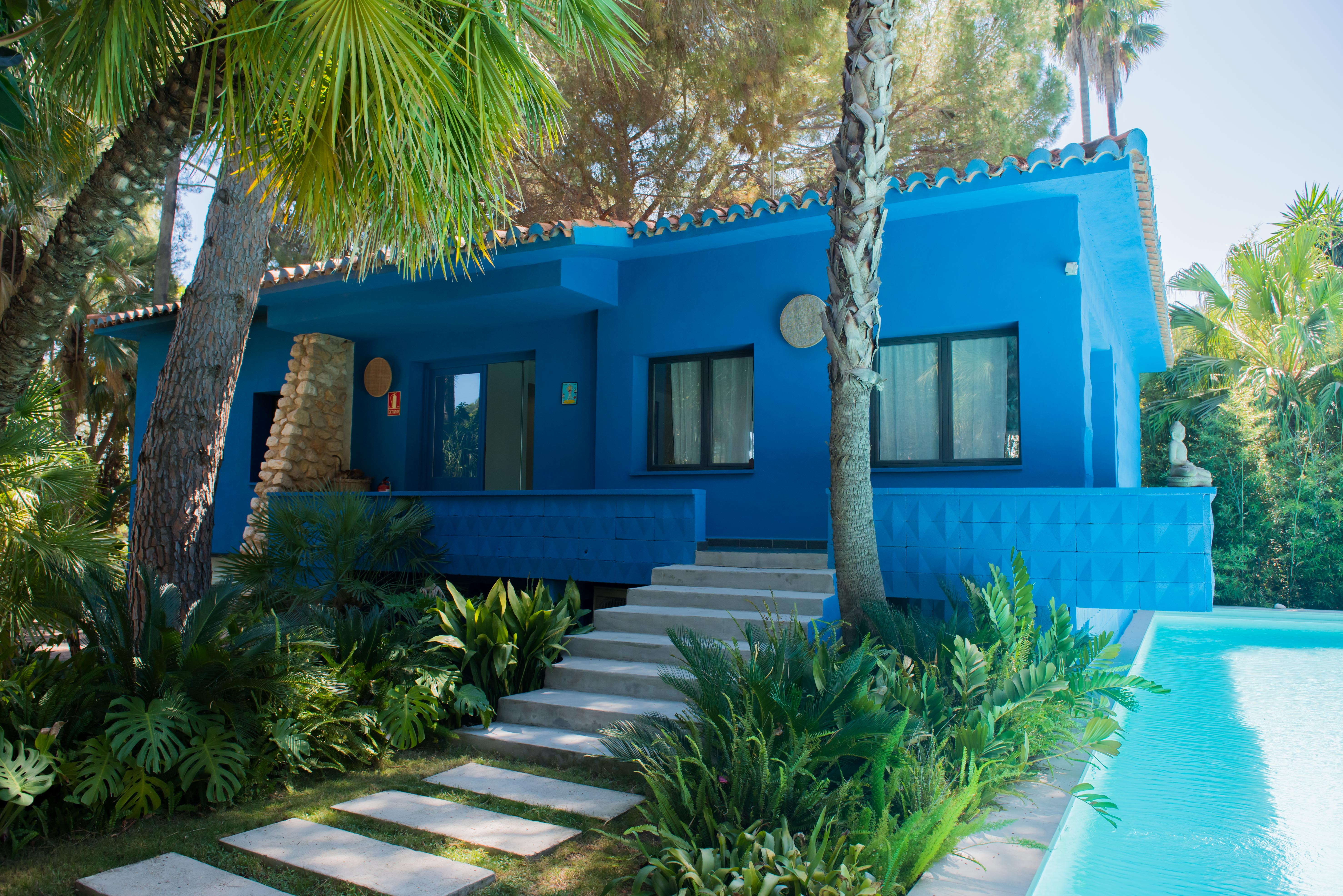 Maison bleu vif avec toit en tuiles, entourée de jardins tropicaux luxuriants et de palmiers. Des escaliers en béton mènent à une terrasse surélevée donnant sur une piscine.