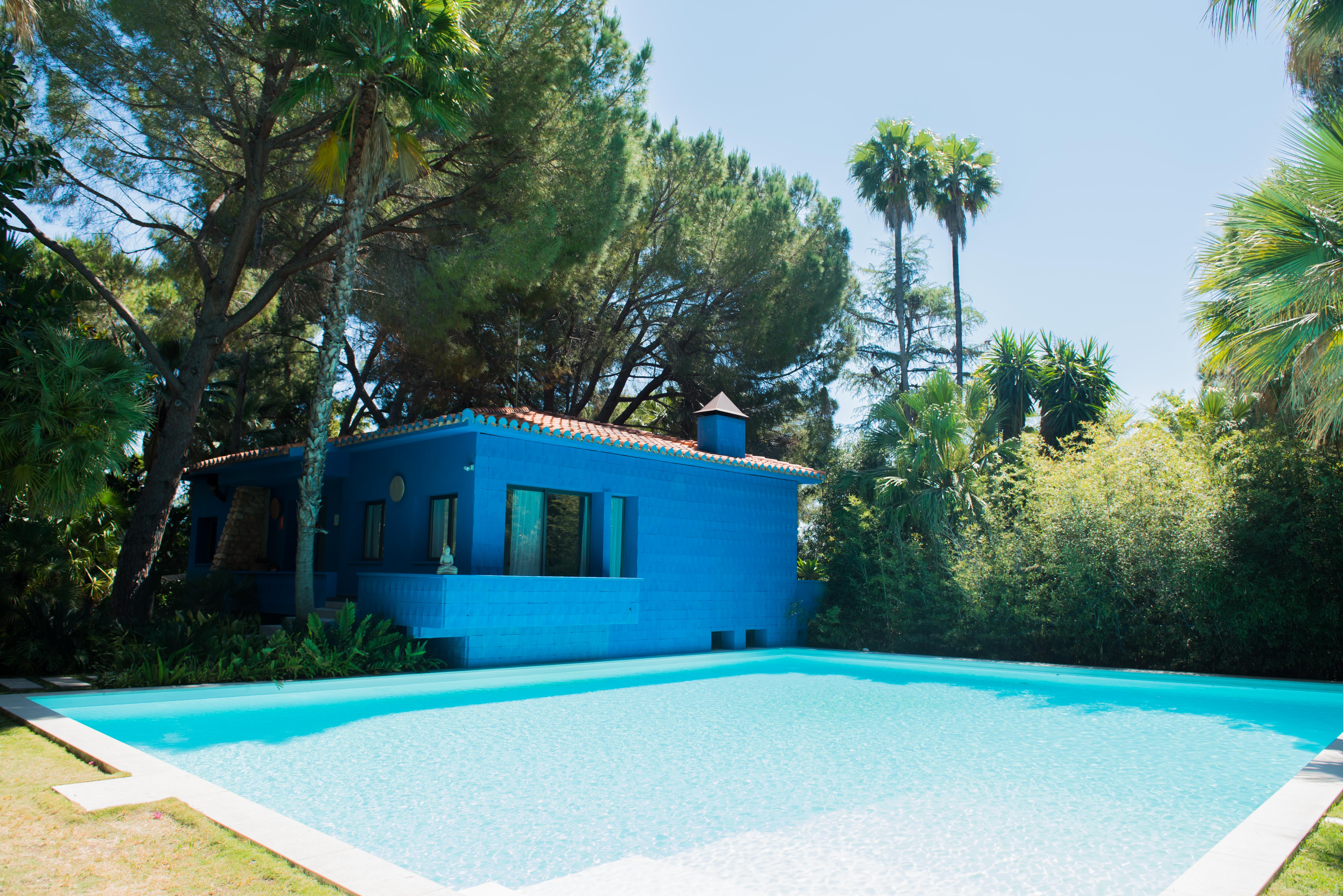 Maison bleue moderne avec toit en terre cuite et piscine sereine bordée d'un dallage clair. Entourée d'une végétation tropicale luxuriante, dont des pins et palmiers, sous un ciel pur.