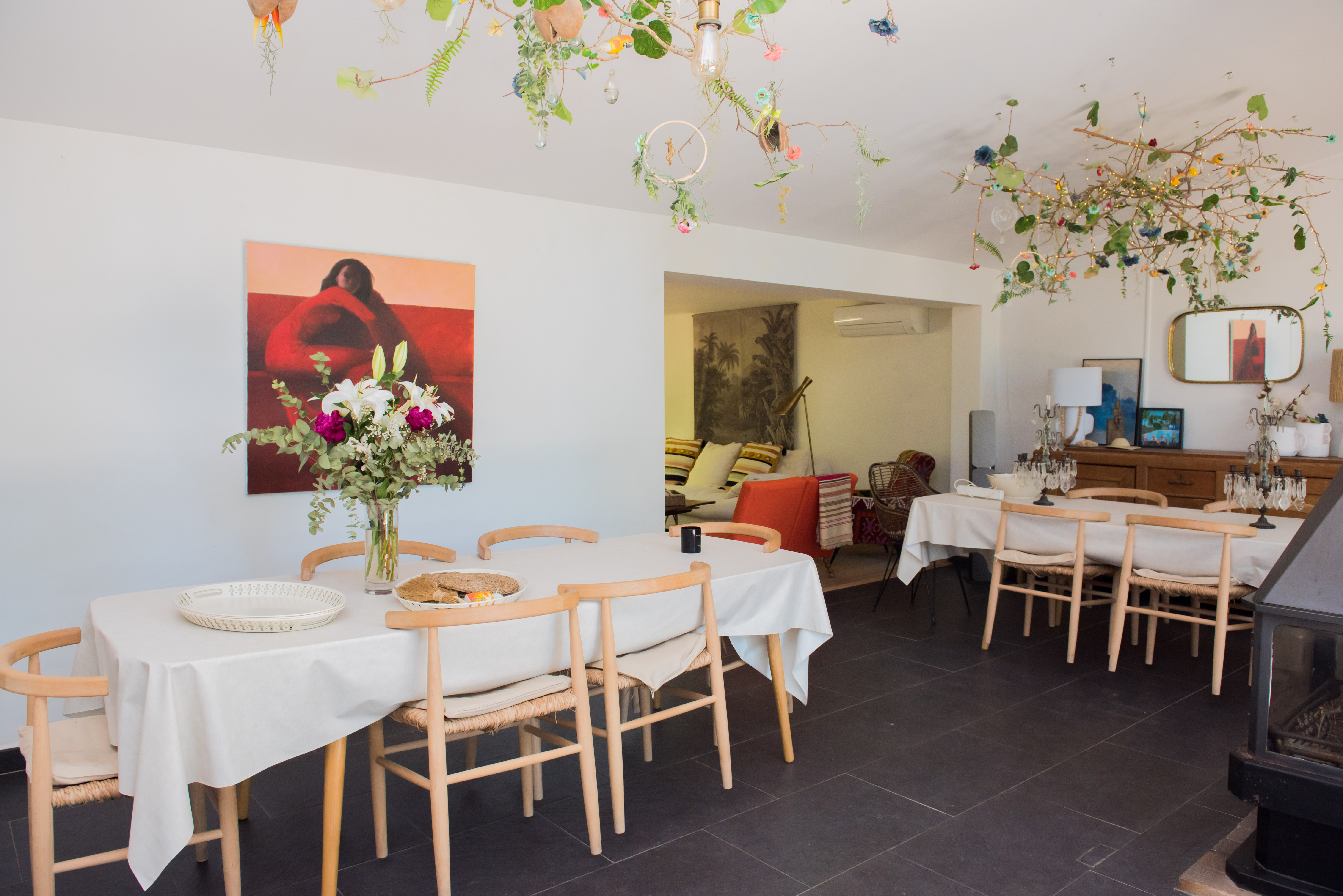 Salle à manger lumineuse avec longue table, chaises en bois clair et arrangement floral frais. Le plafond présente une installation naturelle unique avec branches, fleurs et lumières discrètes, sur un sol carrelé foncé.