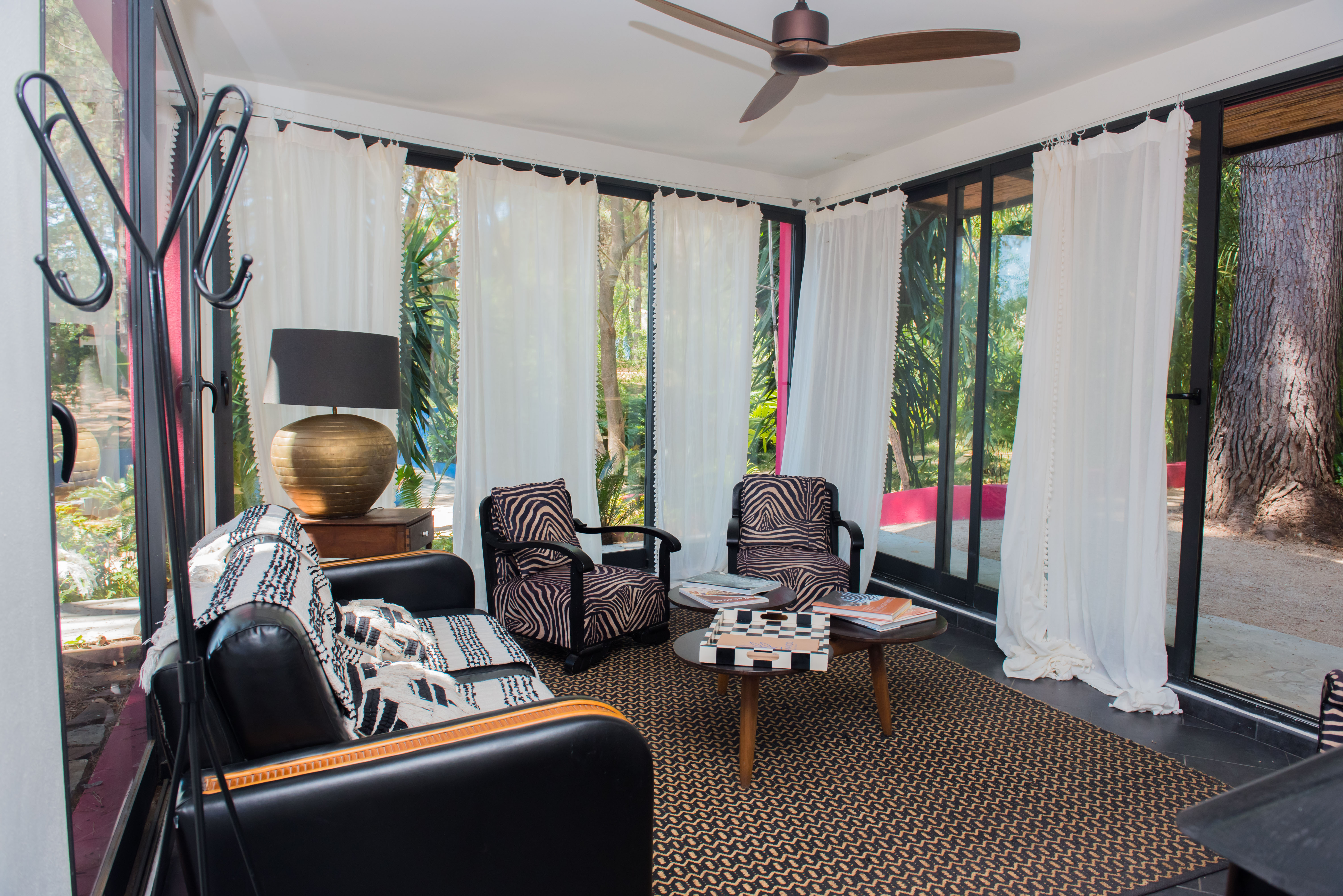 Salon accueillant avec de grandes fenêtres sur un jardin tropical. Il dispose d'un canapé en cuir noir, de fauteuils à imprimé zèbre, d'un tapis géométrique et d'un ventilateur de plafond.