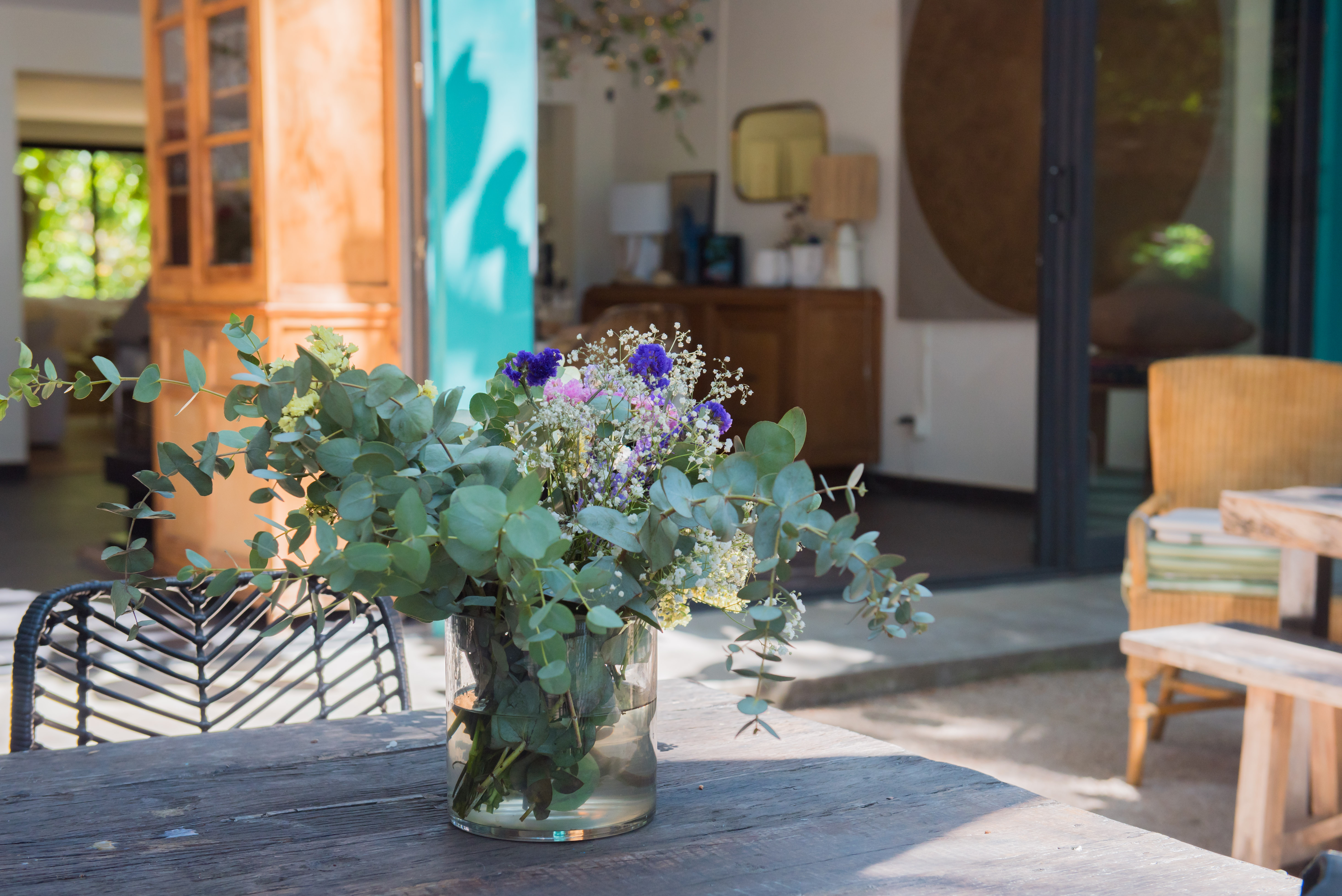 Table extérieure rustique en bois ornée d'un arrangement floral frais d'eucalyptus, avec en arrière-plan une porte turquoise ouverte vers un intérieur lumineux et un jardin verdoyant.