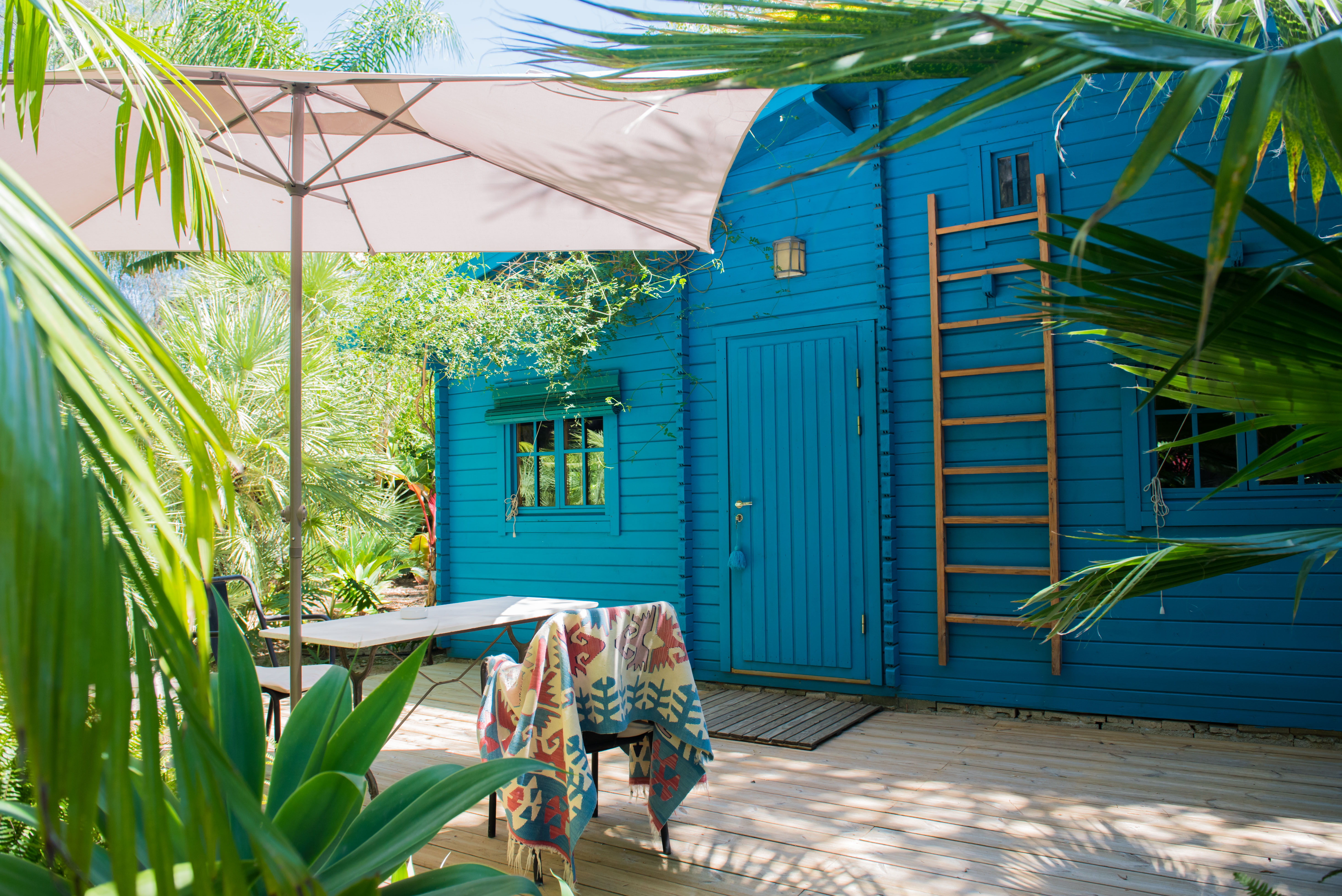 Terrasse en bois avec parasol et mobilier de jardin, jouxtant une cabane bleue éclatante avec échelle en bois, au cœur d'un jardin tropical luxuriant.