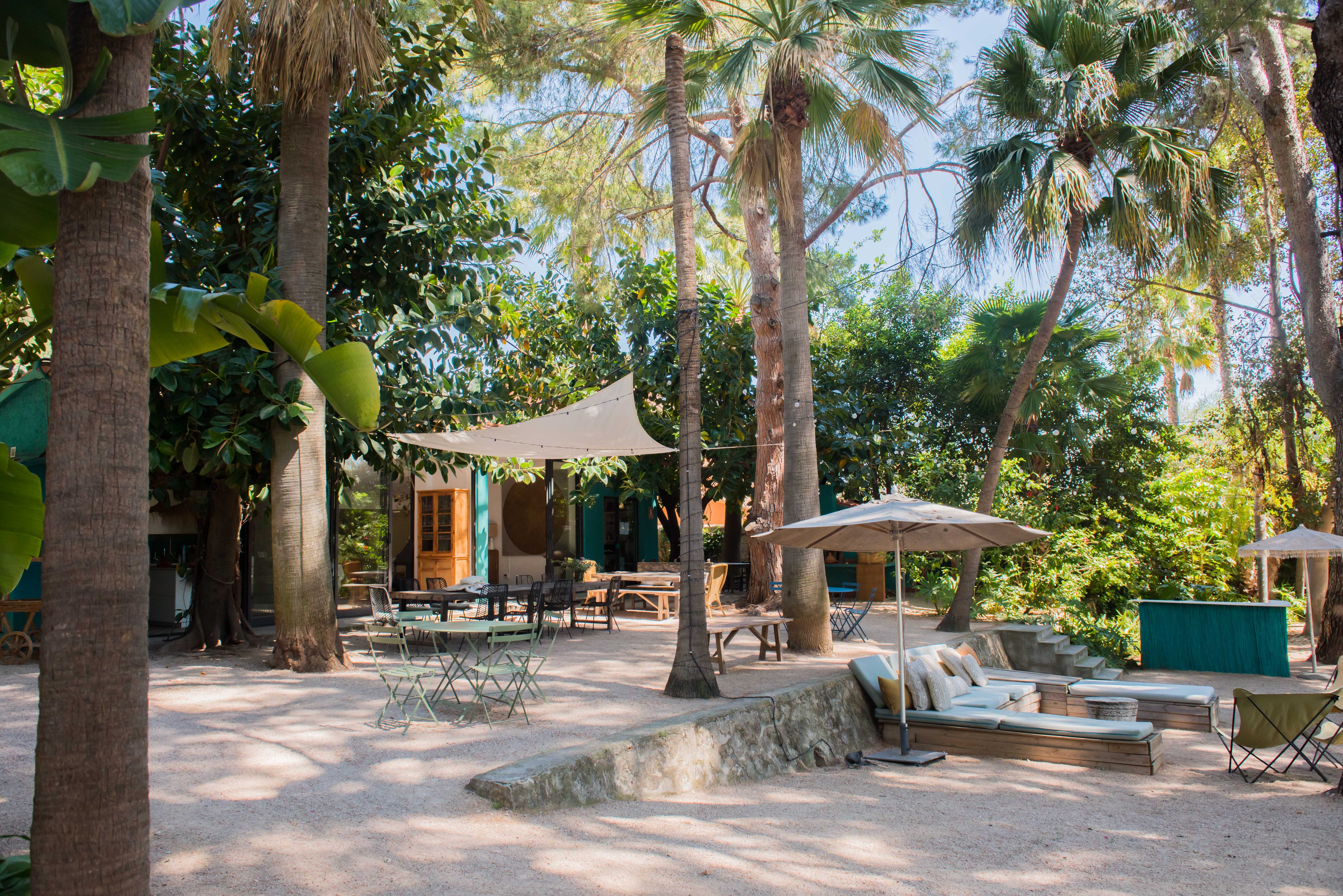 Vaste patio jardin tropical offrant plusieurs espaces de sièges ombragés sur gravier, dont une grande table à manger sous une voile avec guirlandes lumineuses et un coin salon rembourré.