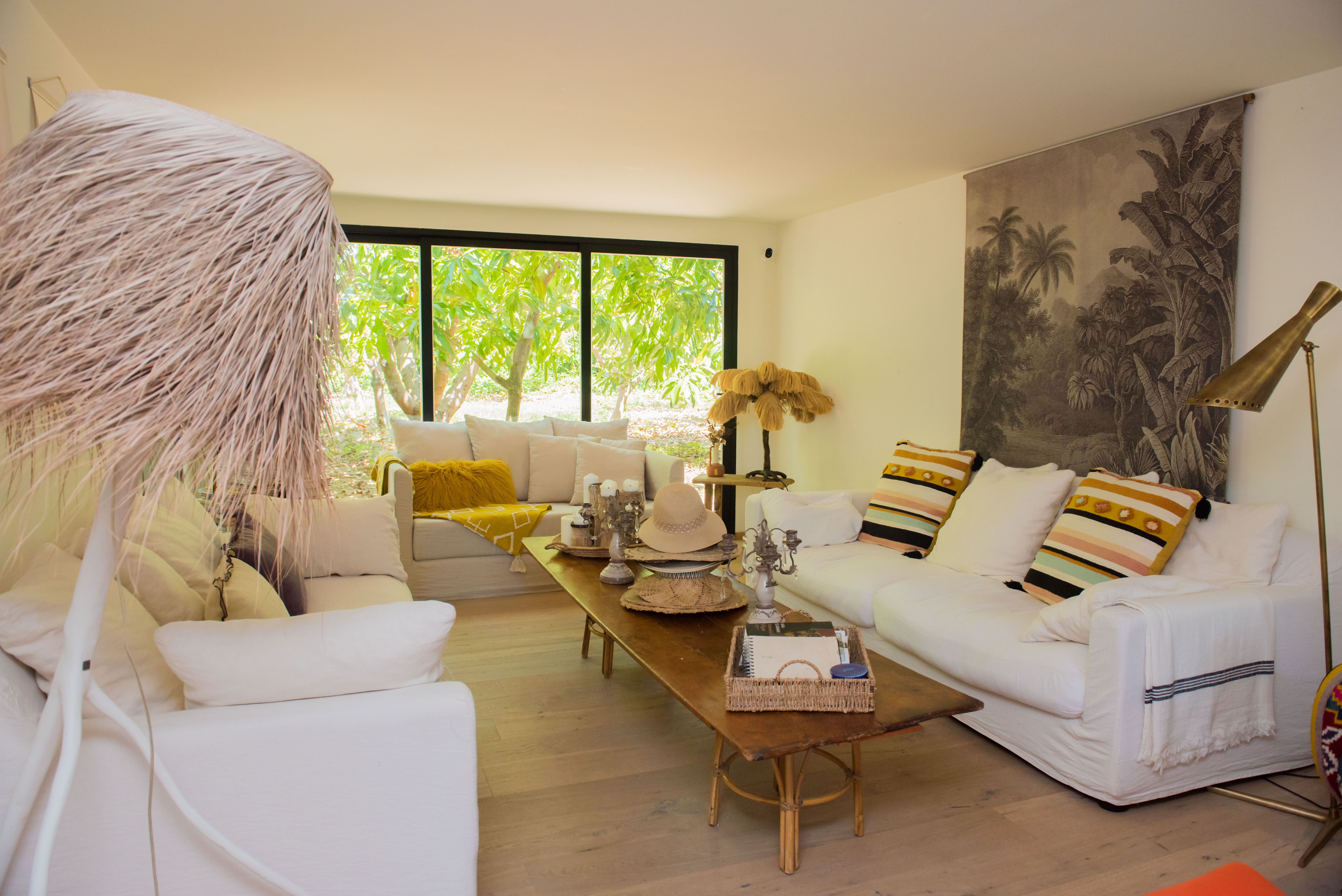 Vaste salon doté d'un parquet clair et de canapés blancs déhoussables. De grandes fenêtres offrent une vue sur un jardin tropical luxuriant, complété par une table basse rustique et des luminaires décoratifs.