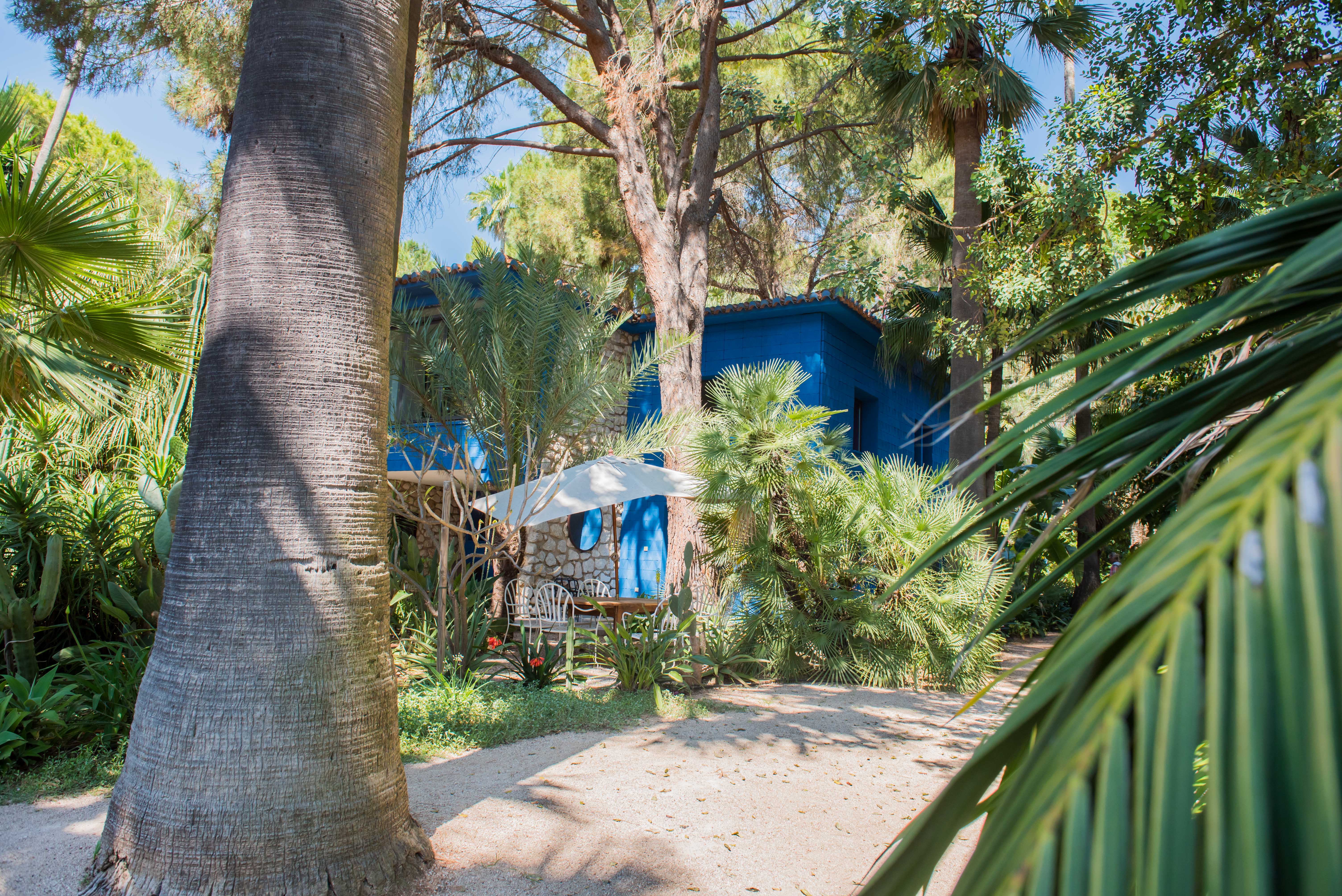 Vue extérieure d'une maison bleue vibrante avec des détails en pierre, nichée dans un jardin tropical luxuriant. Un coin salon extérieur avec des chaises blanches et un parasol est visible sur un chemin de gravier.