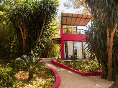 Bâtiment moderne fuchsia avec grandes portes vitrées, auvent en matériaux naturels et balcon supérieur avec sièges en rotin. Un chemin de gravier serpente à travers la végétation tropicale luxuriante.