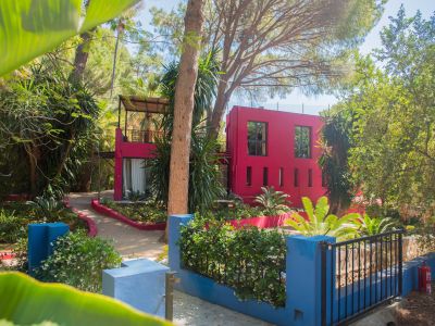 Bâtiment moderne fuchsia éclatant avec fenêtres variées, niché dans un jardin tropical dense avec de grands arbres et des sentiers sinueux. Un muret bleu fleuri délimite le premier plan.