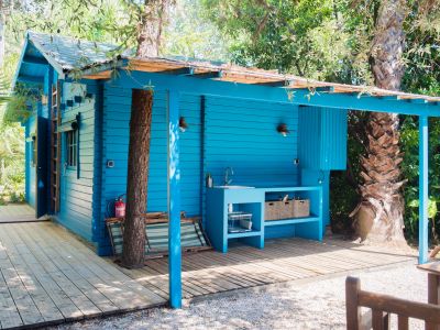 Cabane en bois bleu vif avec cuisine extérieure intégrée, évier et étagères ouvertes, sur une terrasse en bois. Des arbres tropicaux traversent sa canopée naturelle, entourée de chemins de gravier.