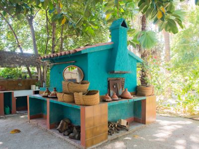 Espace de cuisson extérieur avec four turquoise et cheminée, comptoirs carrelés en terre cuite et rangement pour le bois, niché dans un jardin tropical luxuriant.