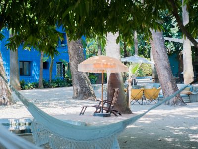 Espace de détente extérieur serein avec un hamac blanc tressé, ombragé par des arbres luxuriants. Un bâtiment bleu texturé et des sièges sous des parasols frangés sont visibles à l'arrière-plan.