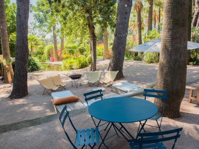 Espace lounge extérieur avec ensemble bistro bleu et chaises en tissu sur gravier, ombragé par des arbres luxuriants avec guirlandes lumineuses, près d'un étang.