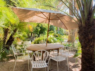 Espace repas extérieur avec table ronde en bois et six chaises en métal blanc sous un grand parasol beige. Entouré de palmiers tropicaux luxuriants et gravier, avec un étang visible en arrière-plan.
