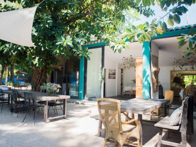 Espace repas extérieur avec tables en bois et sièges variés sous une voile d'ombrage blanche, encadré par une végétation tropicale luxuriante et relié à l'intérieur par des portes vitrées.