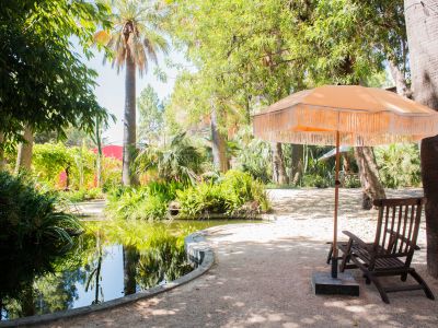 Étang paisible bordé d'une végétation tropicale luxuriante, avec un parasol orange à franges et une chaise longue en bois sur une allée de gravier.