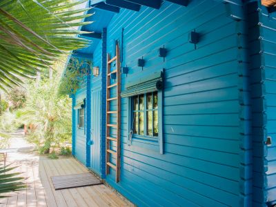 Extérieur d'une cabane en bois bleu, avec échelle décorative et fenêtre à store bambou enroulé. Une terrasse en bois clair s'intègre dans un jardin tropical luxuriant.