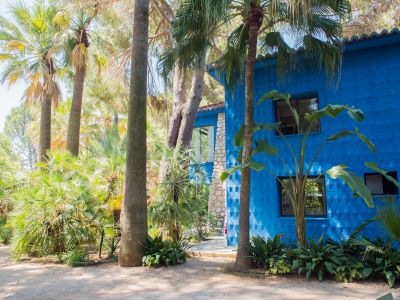 Façade bleue vibrante et texturée d'un bâtiment, entourée d'une végétation tropicale luxuriante. Fenêtres sombres et un toit de tuiles en terre cuite, avec une section en pierre apparente.