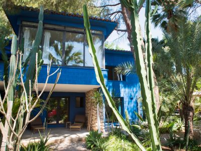 La maison bleue à deux étages offre un patio ombragé au rez-de-chaussée avec chaises longues en bambou et piliers en pierre. Le jardin tropical est encadré par des cactus et palmiers.