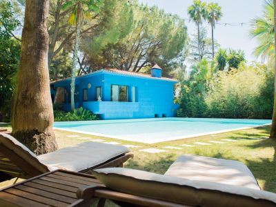 Maison bleu vif à toit de tuiles, bordant une piscine claire. Deux chaises longues en bois sur la pelouse soignée, encadrées par des palmiers et une végétation luxuriante sous des guirlandes lumineuses.