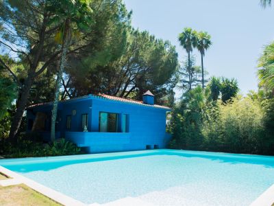 Maison bleue moderne avec toit en terre cuite et piscine sereine bordée d'un dallage clair. Entourée d'une végétation tropicale luxuriante, dont des pins et palmiers, sous un ciel pur.