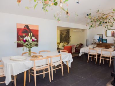Salle à manger lumineuse avec longue table, chaises en bois clair et arrangement floral frais. Le plafond présente une installation naturelle unique avec branches, fleurs et lumières discrètes, sur un sol carrelé foncé.