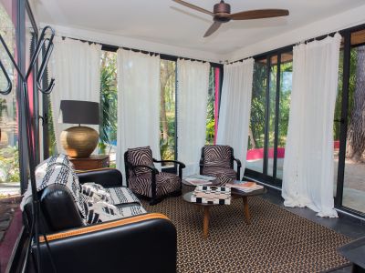 Salon accueillant avec de grandes fenêtres sur un jardin tropical. Il dispose d'un canapé en cuir noir, de fauteuils à imprimé zèbre, d'un tapis géométrique et d'un ventilateur de plafond.