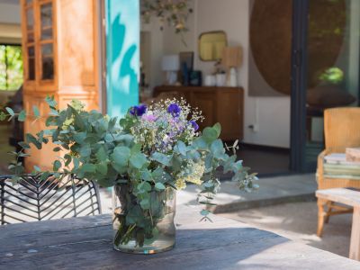 Table extérieure rustique en bois ornée d'un arrangement floral frais d'eucalyptus, avec en arrière-plan une porte turquoise ouverte vers un intérieur lumineux et un jardin verdoyant.