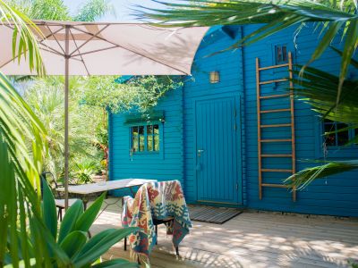 Terrasse en bois avec parasol et mobilier de jardin, jouxtant une cabane bleue éclatante avec échelle en bois, au cœur d'un jardin tropical luxuriant.