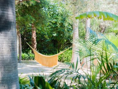 Un hamac jaune à franges offre un lieu de détente serein, niché entre deux palmiers dans un jardin tropical ensoleillé avec chemin de gravier clair.