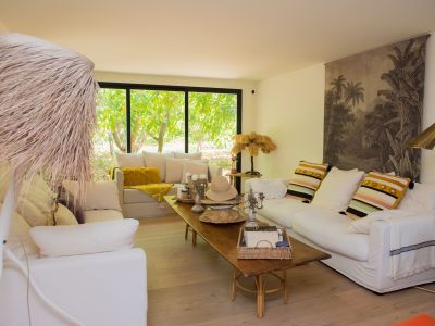 Vaste salon doté d'un parquet clair et de canapés blancs déhoussables. De grandes fenêtres offrent une vue sur un jardin tropical luxuriant, complété par une table basse rustique et des luminaires décoratifs.