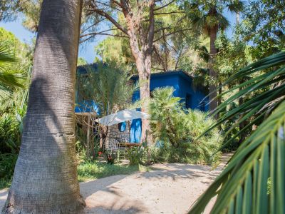 Vue extérieure d'une maison bleue vibrante avec des détails en pierre, nichée dans un jardin tropical luxuriant. Un coin salon extérieur avec des chaises blanches et un parasol est visible sur un chemin de gravier.