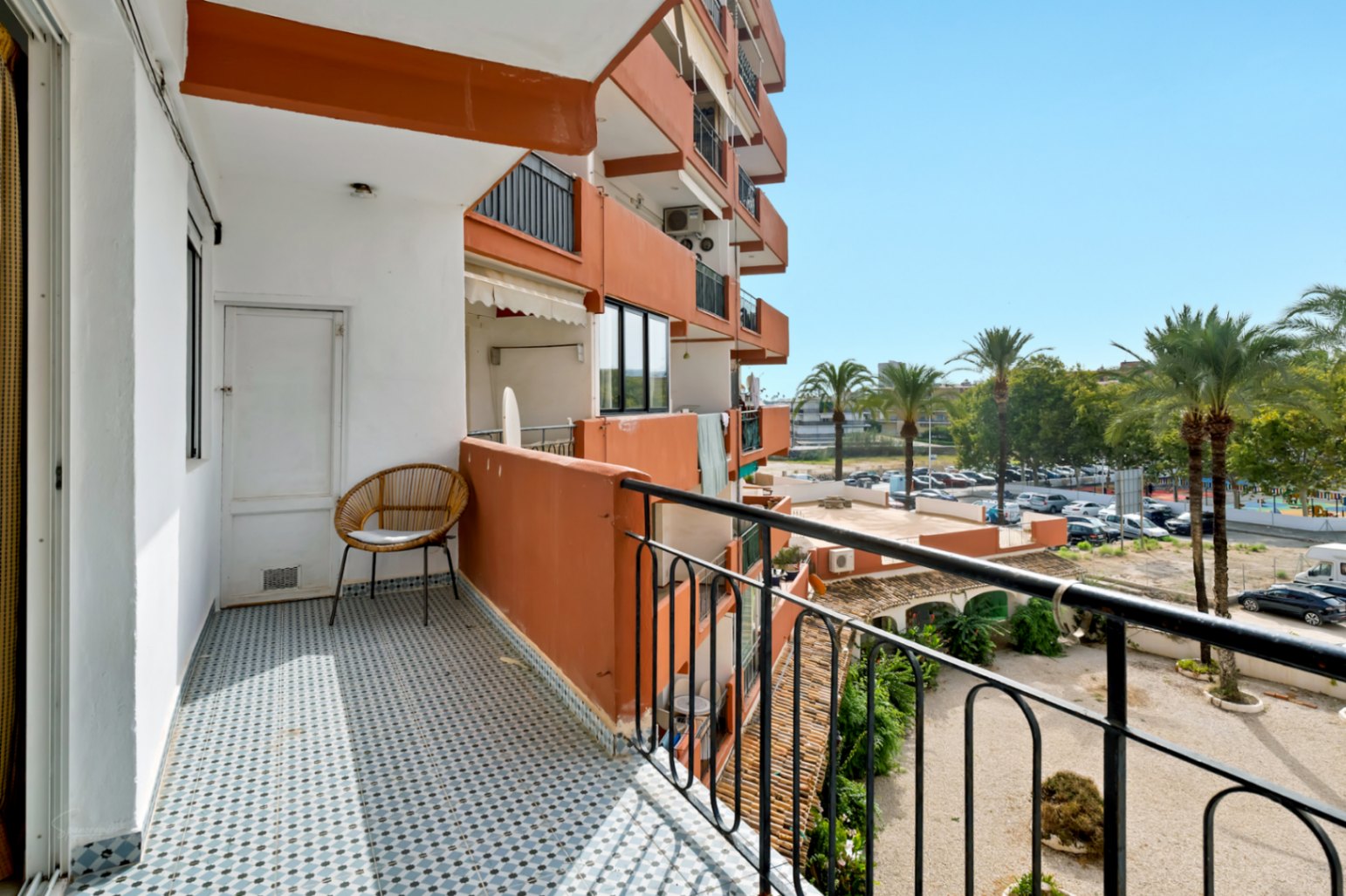 Balcon ensoleillé avec carrelage géométrique bleu et blanc, doté d'une chaise en osier et d'une porte de rangement. Vues sur les palmiers et le parking résidentiel.
