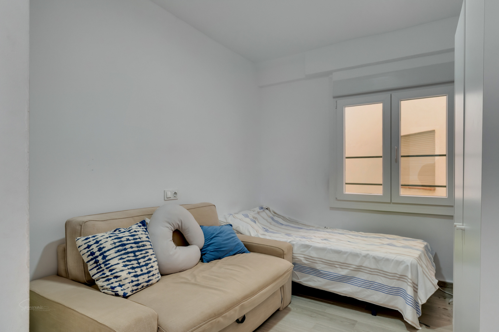 Chambre d'appoint avec canapé-lit beige et lit simple, murs blancs et fenêtre apportant une lumière naturelle diffuse.