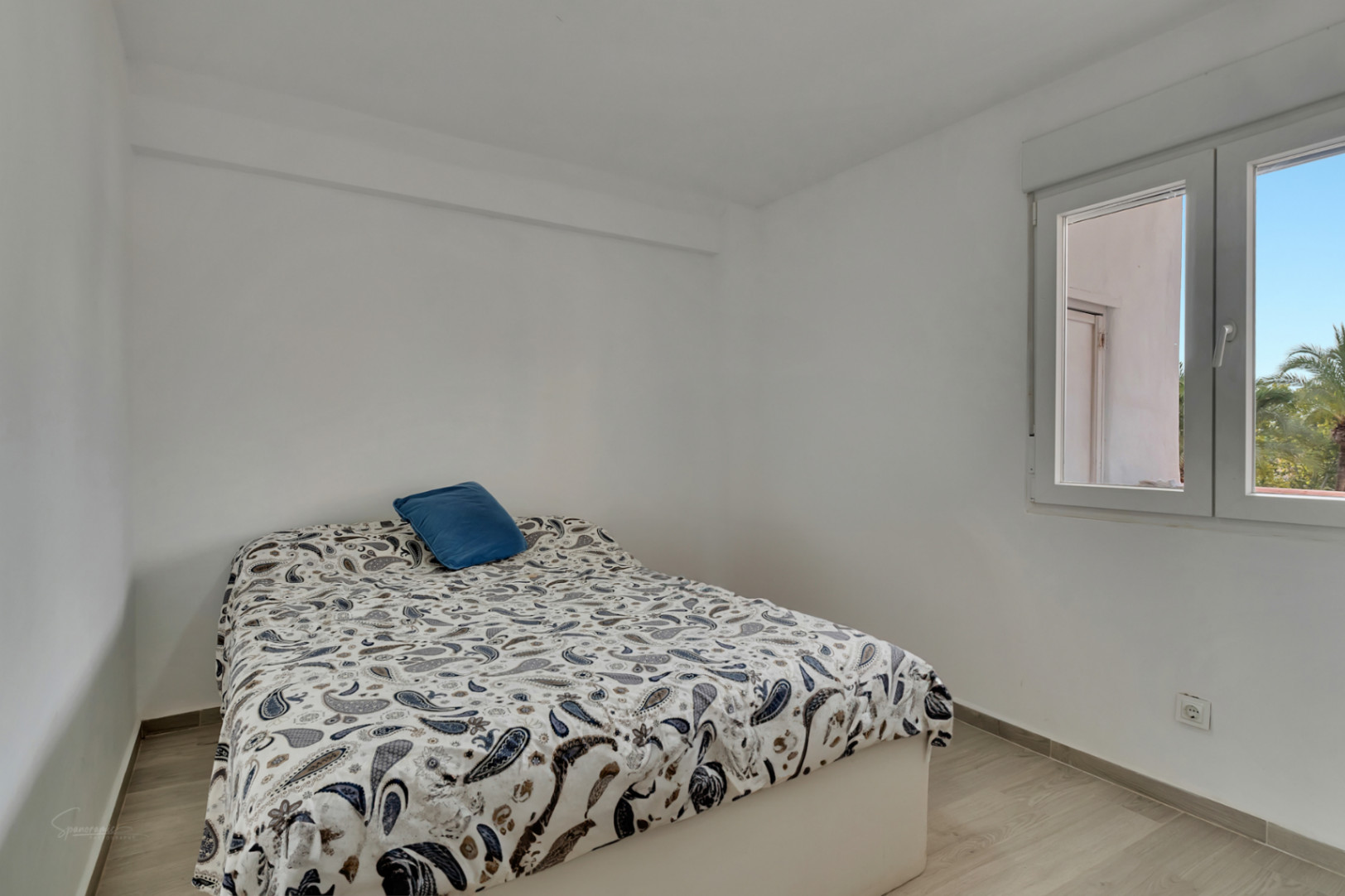 Chambre lumineuse aux murs blancs et sol stratifié clair. Un lit avec une couette à motif cachemire et un oreiller bleu est placé près d'une fenêtre, offrant lumière naturelle et vue sur des palmiers.
