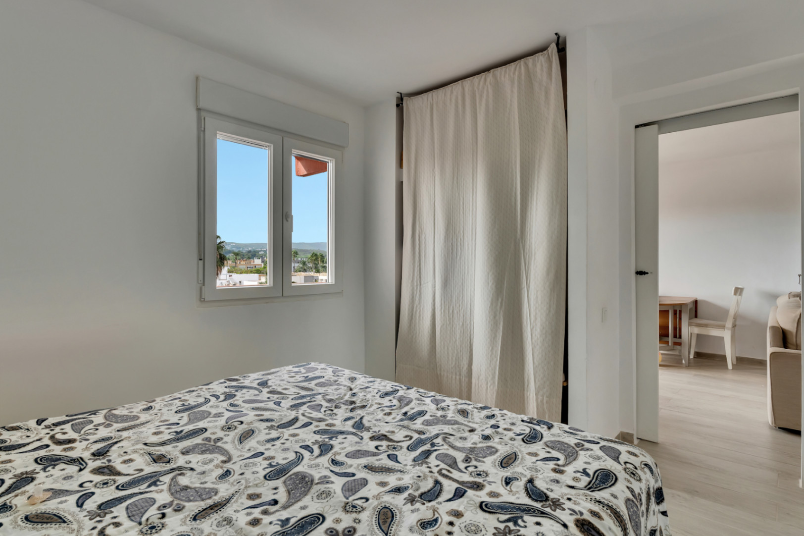 Chambre lumineuse avec lit à motifs, fenêtre offrant des vues urbaines et une armoire discrète couverte par un rideau clair, avec accès à une pièce adjacente.