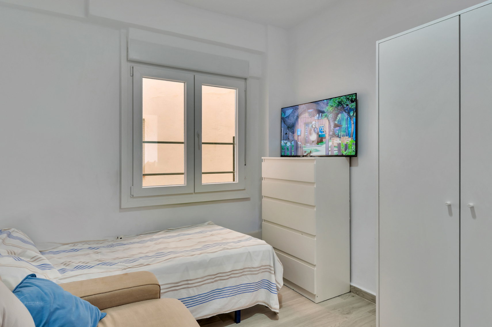 Chambre secondaire avec un lit simple, une armoire intégrée blanche et une commode surmontée d'une télévision. La fenêtre à double battant apporte de la lumière naturelle.