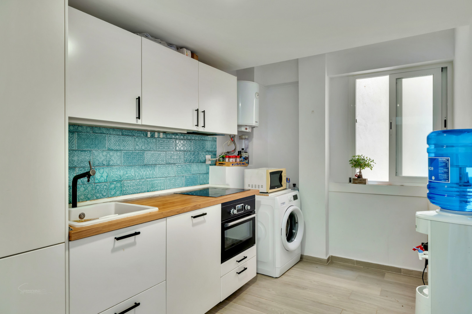 Cuisine moderne avec armoires blanches, plan de travail en bois clair et crédence en carreaux turquoise à motifs. Équipée d'appareils intégrés et d'un espace buanderie avec lave-linge.