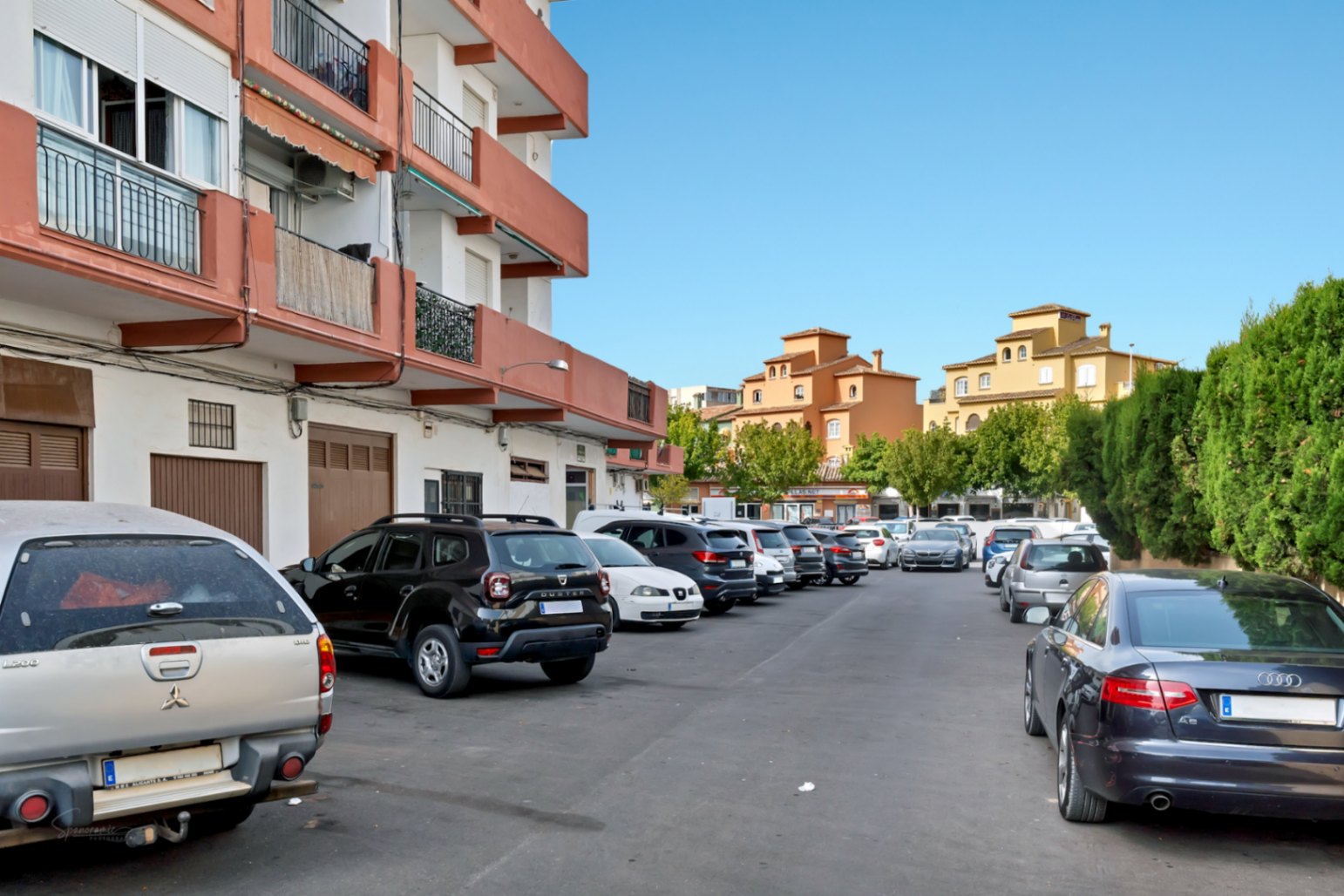 Espace de stationnement extérieur avec plusieurs véhicules, bordé par un immeuble résidentiel aux balcons orange et des structures ocre traditionnelles en arrière-plan sous un ciel clair.