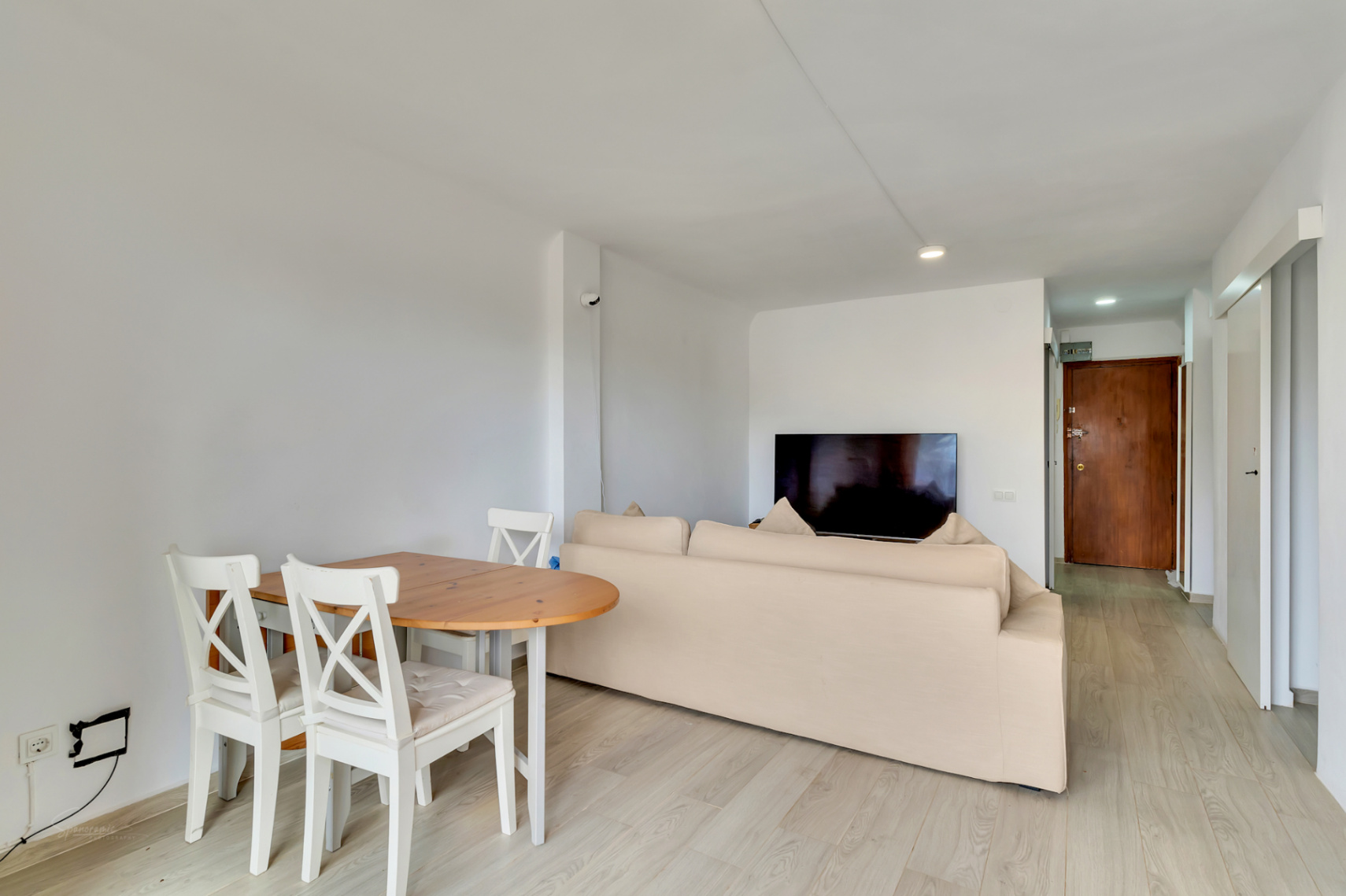 Espace salon-salle à manger ouvert avec table en bois clair, chaises blanches et canapé beige. Télévision murale et sol stratifié clair menant à une porte d'entrée en bois foncé.