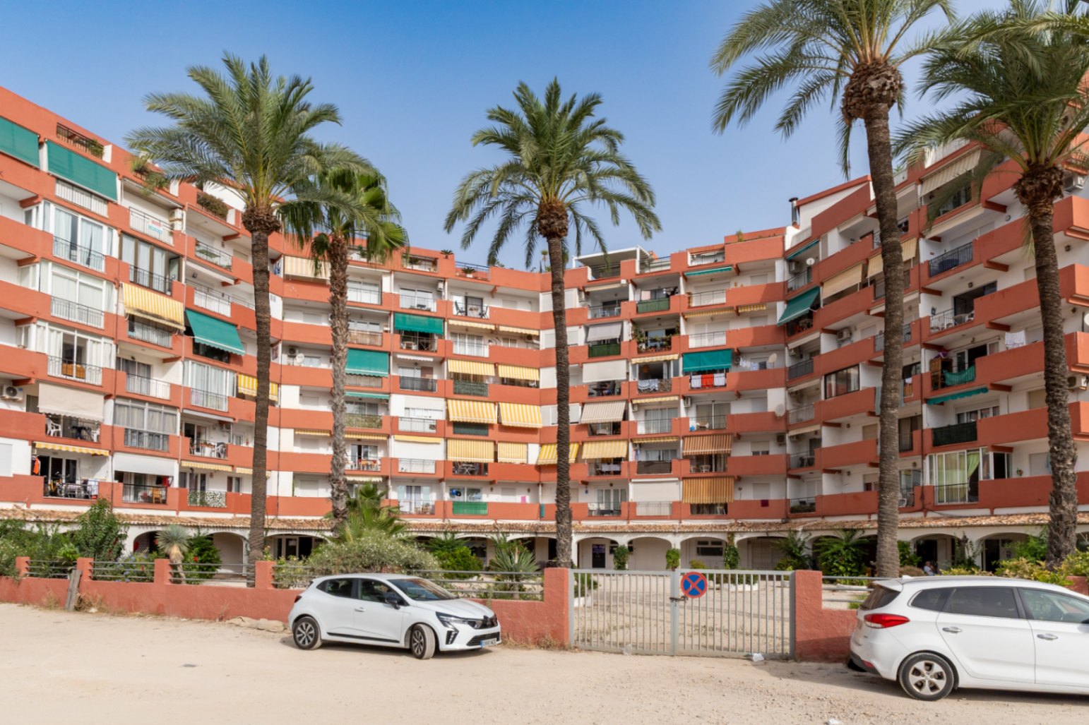 Immeuble d'appartements incurvé avec façade terre cuite et balcons aux stores variés. De grands palmiers en premier plan, avec un parking extérieur et un portail.