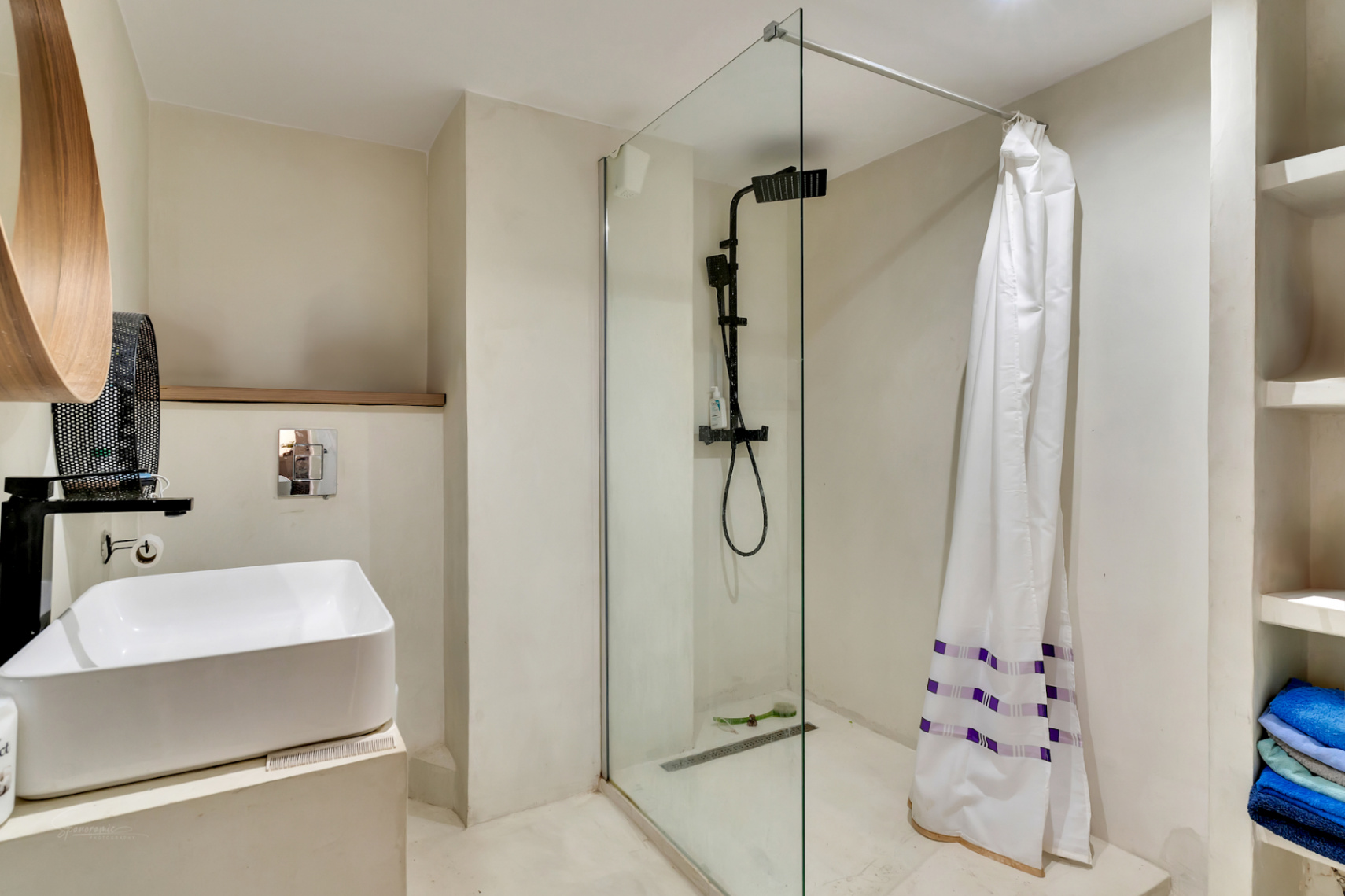 Salle de bain moderne avec lavabo vasque, robinetterie noire mate et douche à l'italienne avec paroi vitrée. Miroir en bois distinctif et étagères intégrées.