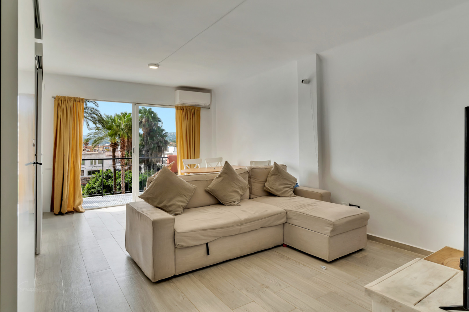 Salon lumineux avec canapé d'angle beige, sol effet bois clair et accès à un balcon offrant des vues sur les palmiers et des rideaux jaunes.