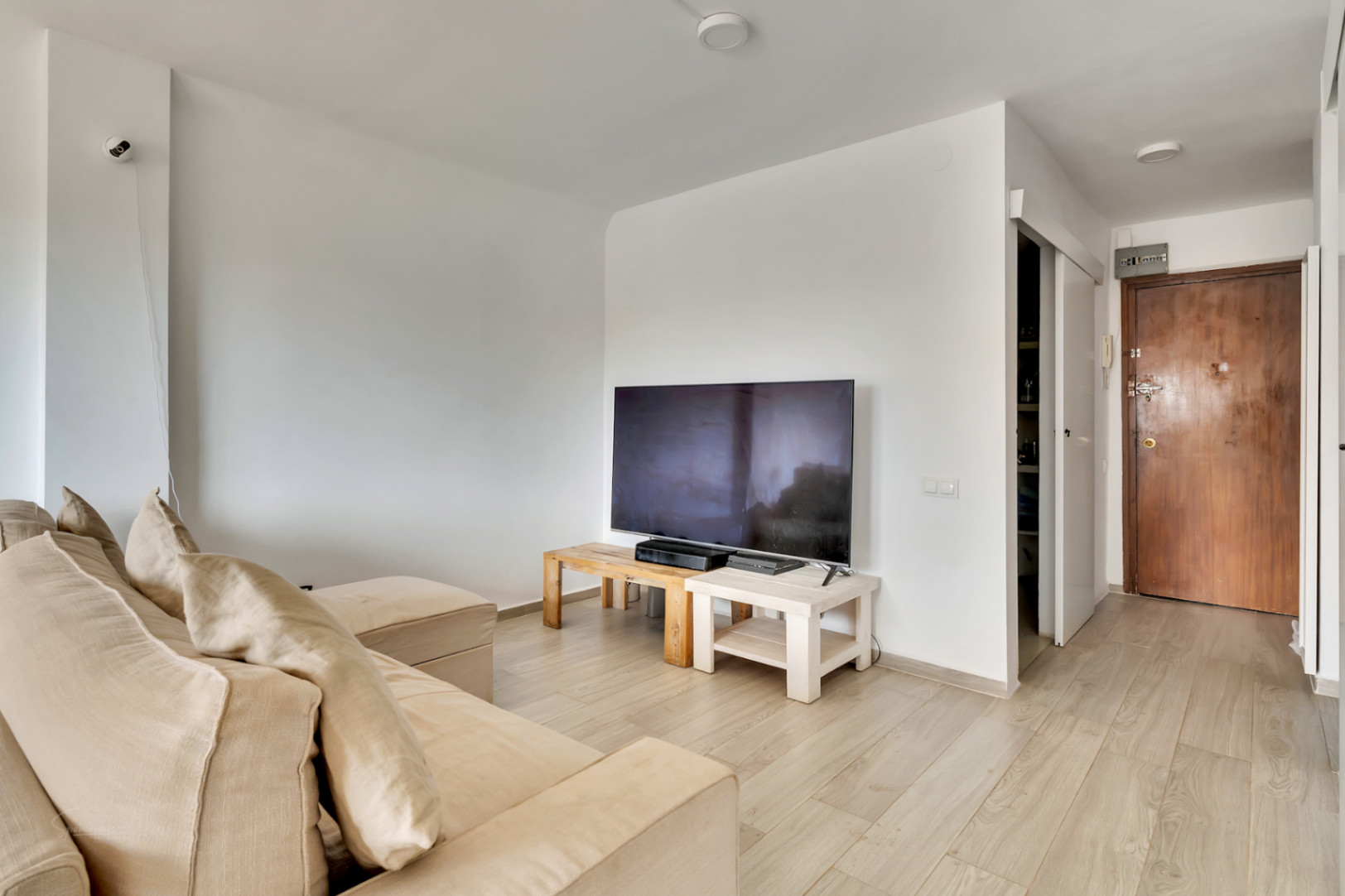 Séjour lumineux avec canapé sectionnel beige et TV sur des tables en bois clair. Le sol stratifié clair mène à la porte d'entrée en bois foncé et à un espace de rangement étagère.