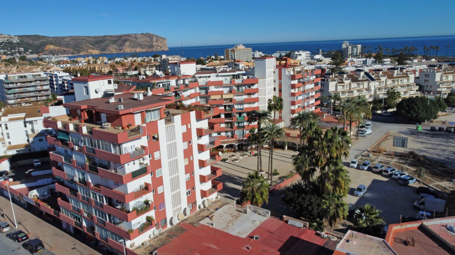 Vue aérienne du complexe résidentiel avec façades blanches, balcons rouge-orange, palmiers et la mer Méditerranée en arrière-plan.