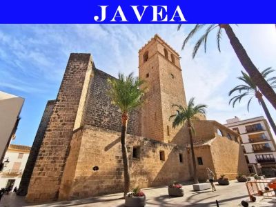 Ancienne église en pierre avec clocher à Jávea, montrant sa maçonnerie robuste et ses contreforts, sur une place ensoleillée bordée de palmiers.