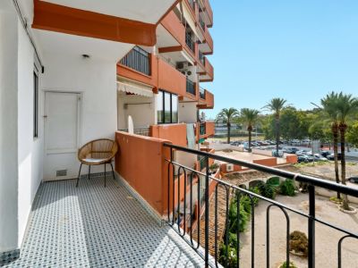Balcon ensoleillé avec carrelage géométrique bleu et blanc, doté d'une chaise en osier et d'une porte de rangement. Vues sur les palmiers et le parking résidentiel.