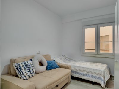 Chambre d'appoint avec canapé-lit beige et lit simple, murs blancs et fenêtre apportant une lumière naturelle diffuse.