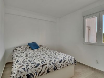 Chambre lumineuse aux murs blancs et sol stratifié clair. Un lit avec une couette à motif cachemire et un oreiller bleu est placé près d'une fenêtre, offrant lumière naturelle et vue sur des palmiers.